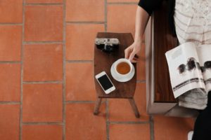 Person in cafe, sitting in chair next to small table with a cup of coffee, cellphone and camera.